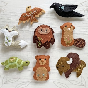 May include: A set of felt animal ornaments, including a fox, a bear, a turtle, a beaver, a bison, a crow, and a bird. The ornaments are all hand-stitched and feature embroidered details.