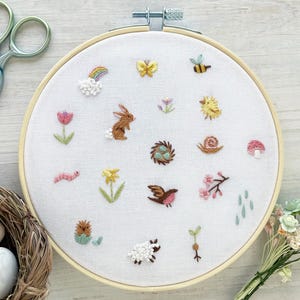 Op de afbeelding: Een witte borduurring met verschillende kleurrijke geborduurde ontwerpen, waaronder een konijn, een vogel, een slak, een paddenstoel, een bloem, een bij, een vlinder en een regenboog. De ring ligt op een licht houten oppervlak.