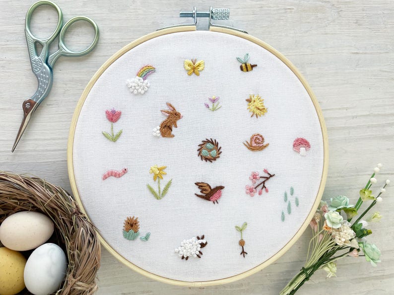 Op de afbeelding: Een witte borduurring met verschillende kleurrijke geborduurde ontwerpen, waaronder een konijn, een vogel, een slak, een paddenstoel, een bloem, een bij, een vlinder en een regenboog. De ring ligt op een licht houten oppervlak.