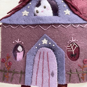 May include: A felt house in shades of purple, with a cat peering from the attic window. The house features a purple roof, a lavender door, and decorative embroidered details like stars, roses, and a spiderweb. A whimsical, handmade item.