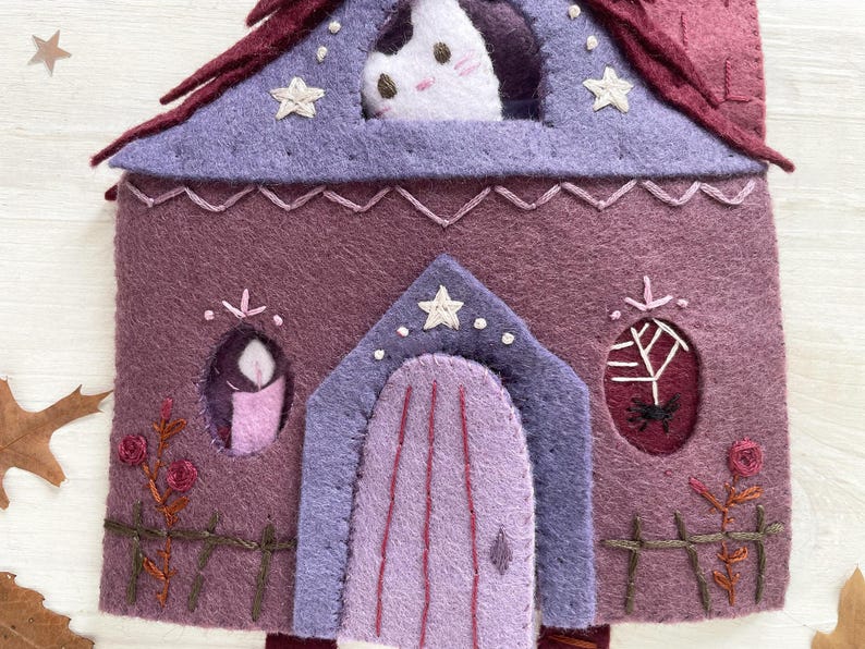 May include: A felt house in shades of purple, with a cat peering from the attic window. The house features a purple roof, a lavender door, and decorative embroidered details like stars, roses, and a spiderweb. A whimsical, handmade item.