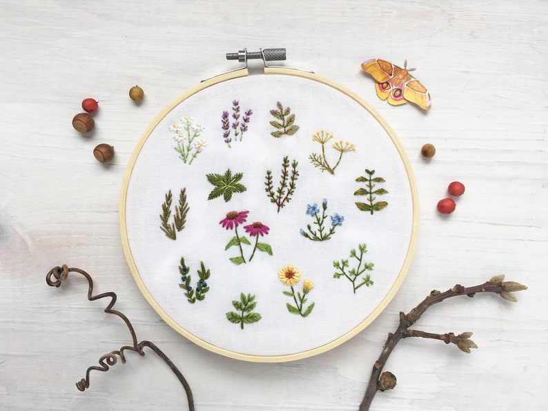 May include: A white embroidery hoop with a variety of floral and leaf designs embroidered on it. The hoop is surrounded by small acorns, berries, and a twig with buds.