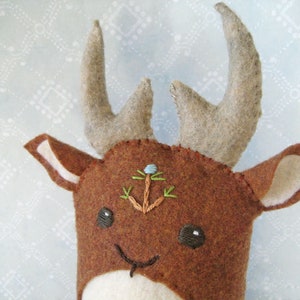May include: A brown felt deer with a white face and large antlers. The deer has embroidered eyes and a small blue flower on its forehead.