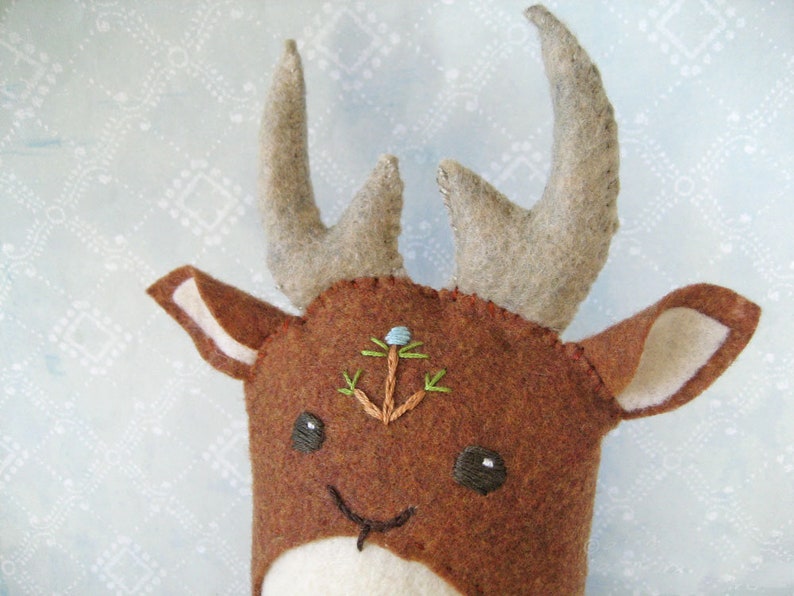 May include: A brown felt deer with a white face and large antlers. The deer has embroidered eyes and a small blue flower on its forehead.