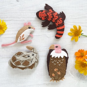 Op de afbeelding: Vijf vilt dierlijke applicaties, waaronder een bruine en witte muis, een bruine schildpad, een rode en zwarte hagedis, een bruine en witte vogel en een gele bloem.