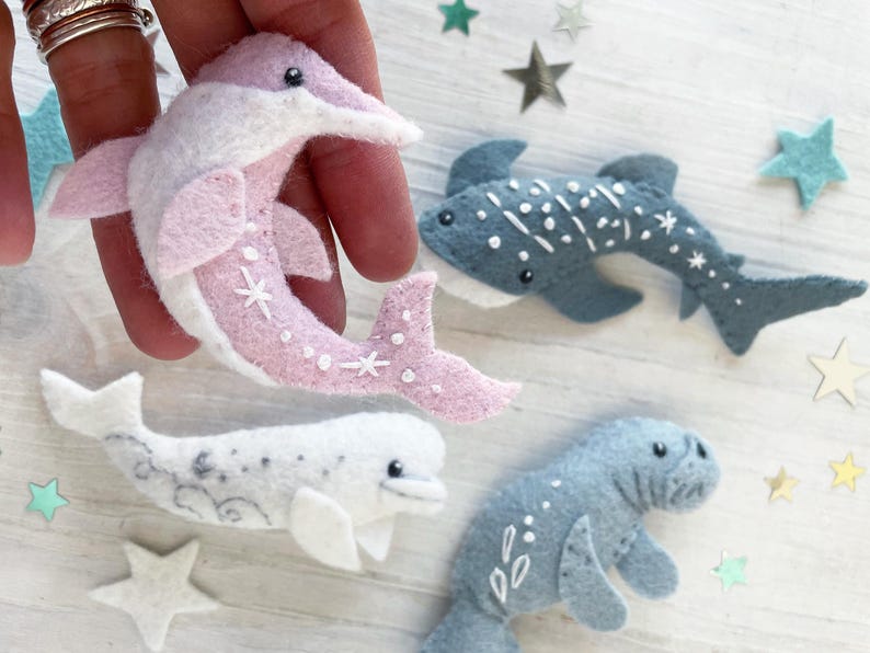 May include: Four felt animal toys: a pink dolphin, a blue whale, a white beluga whale, and a gray shark. Each toy has a white star pattern.