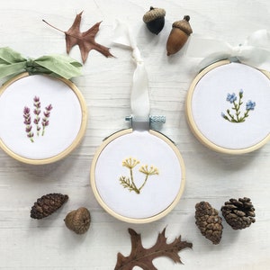 May include: Three embroidery hoops with floral designs. The hoops are made of wood and have a white fabric background. The designs are of lavender, forget-me-nots, and a yellow wildflower.