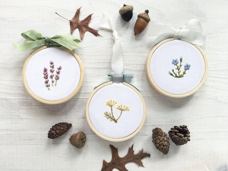May include: Three embroidery hoops with floral designs. The hoops are made of wood and have a white fabric background. The designs are of lavender, forget-me-nots, and a yellow wildflower.