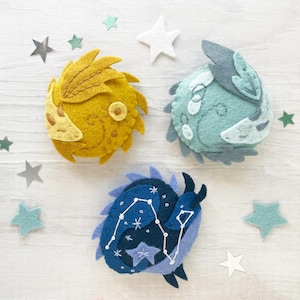 Op de afbeelding: Drie vilten ornamenten in de vorm van hemellichamen. De eerste is een gele zon met een gezicht en stralen. De tweede is een blauwe en groene maan met een gezicht en kraters. De derde is een blauwe en witte constellatie met een ster in het midden.