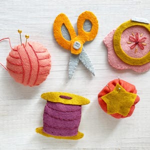 May include: A collection of handmade felt sewing accessories, including a yarn ball with pins, orange scissors, an embroidery hoop, a spool of thread, and a pincushion with a star. The items are in shades of orange, pink, yellow, and purple.