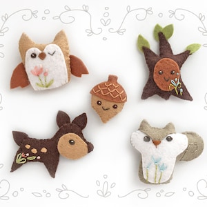 May include: Five felt woodland animal ornaments: a brown owl with pink flowers, a brown acorn with a smiling face, a brown tree with green leaves, a brown deer with white spots, and a gray squirrel with blue flowers.