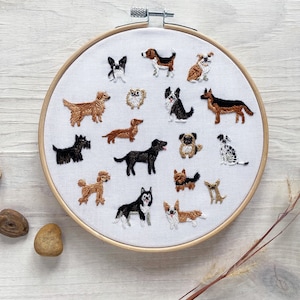 May include: Embroidered hoop art featuring various dog breeds. The white fabric is adorned with detailed, colorful thread depictions of dogs, including a Dalmatian, Poodle, and Dachshund. The wooden hoop is displayed on a light-colored wooden surface.