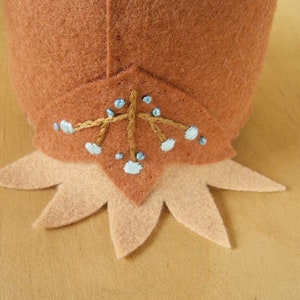 Pu&ograve; includere: Un cappello da gnomo in feltro marrone con un bordo in feltro marrone chiaro e un motivo ricamato decorativo di fiori blu e dorati.