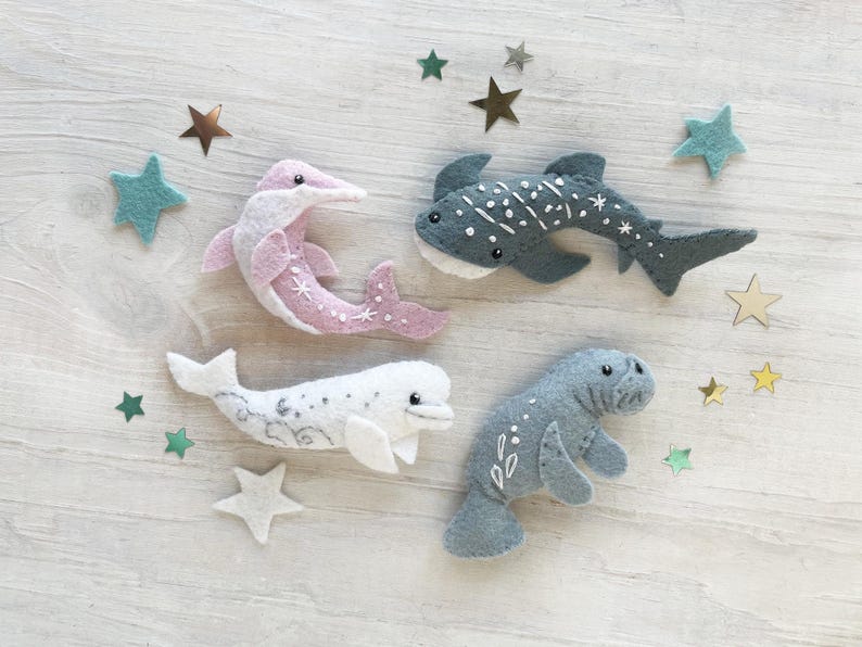 May include: Four felt animal toys: a pink dolphin, a gray whale shark, a white beluga whale, and a gray manatee. Each toy has embroidered details and a whimsical design.