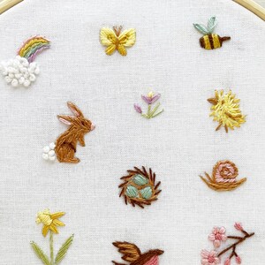 Op de afbeelding: Borduurwerk op een borduurring met verschillende lentethema's, waaronder een konijn, een vlinder, een bij, een slak, een paddenstoel, een bloem, een vogel, een nest en een worm.