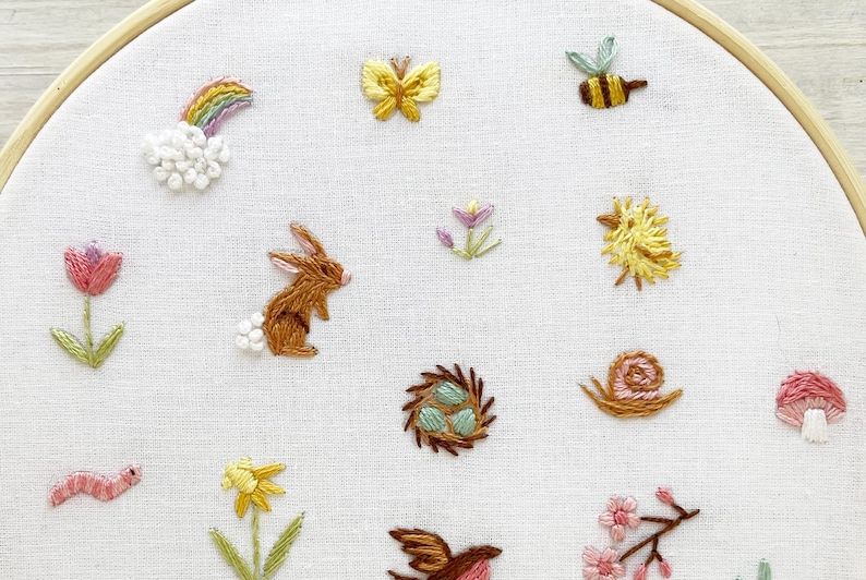 Op de afbeelding: Borduurwerk op een borduurring met verschillende lentethema's, waaronder een konijn, een vlinder, een bij, een slak, een paddenstoel, een bloem, een vogel, een nest en een worm.
