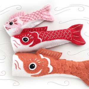 May include: Three felt fish ornaments in pink, red, and orange with white bellies and embroidered details. The fish are hanging from a white string.