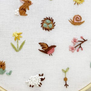 Op de afbeelding: Een witte borduurring met verschillende kleurrijke geborduurde ontwerpen, waaronder een vogel, een slak, een bloem, een konijn, een paddenstoel, een nest, een worm, een egel, een scheut en een schaap.
