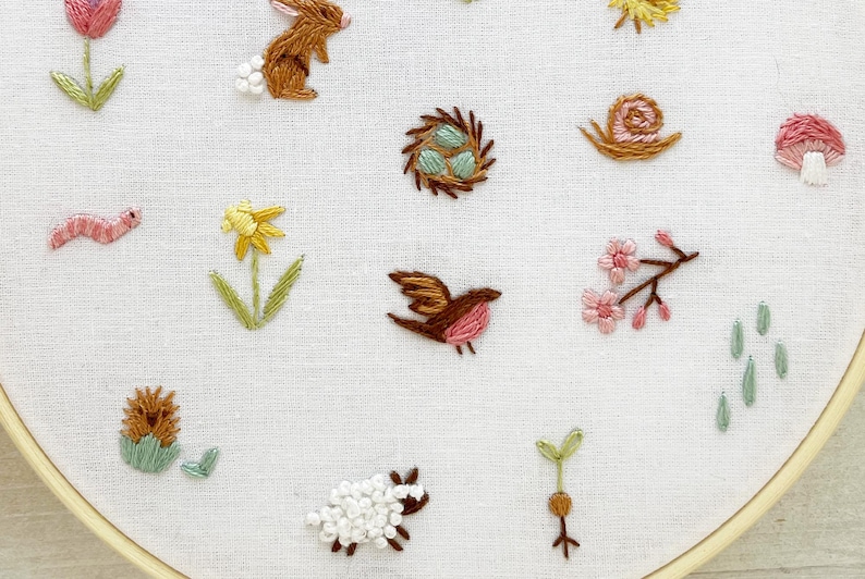 Op de afbeelding: Een witte borduurring met verschillende kleurrijke geborduurde ontwerpen, waaronder een vogel, een slak, een bloem, een konijn, een paddenstoel, een nest, een worm, een egel, een scheut en een schaap.