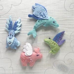 May include: A collection of four felt creatures: a blue winged dolphin, a blue and white winged rabbit, a pink pig with white wings, and a green frog with purple wings. The creatures are handmade and have embroidered details.