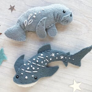 May include: Two felt animal toys, a gray manatee and a blue whale shark, with white embroidered details. The toys are on a white wooden background.