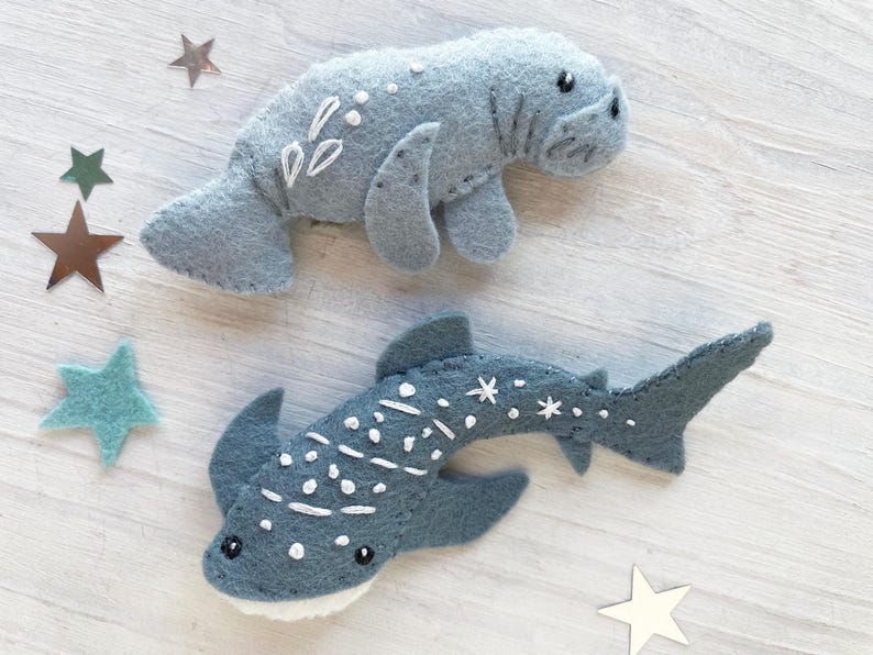 May include: Two felt animal toys, a gray manatee and a blue whale shark, with white embroidered details. The toys are on a white wooden background.