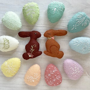 May include: A set of felt Easter eggs and bunnies with embroidered floral designs. The eggs are in various pastel colors, including light blue, green, yellow, pink, and white. The bunnies are brown with floral embroidery.