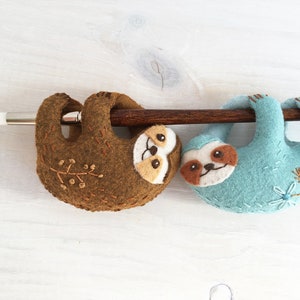 May include: Two felt sloth ornaments hanging from a wooden branch. The sloth on the left is brown with a light brown face and the sloth on the right is light blue with a light brown face.