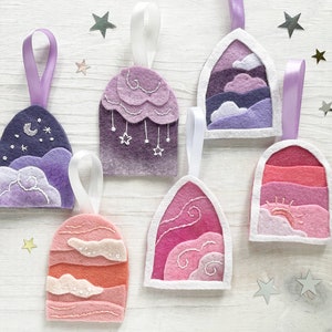 Pode incluir: Seis enfeites de feltro com desenhos bordados de nuvens, estrelas e luas. Os enfeites são em tons de rosa, roxo e branco e estão pendurados com uma fita branca.