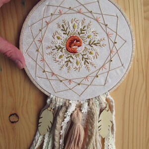 May include: Embroidered dreamcatcher featuring a central design of a sleeping fox in orange thread, surrounded by floral and geometric patterns. The hoop is adorned with beige and cream-colored yarn, lace, and a tassel, with two leaf-shaped pendants.