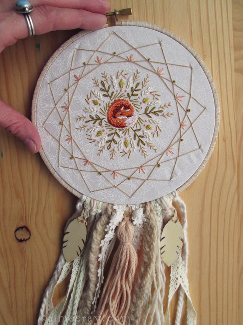 May include: Embroidered dreamcatcher featuring a central design of a sleeping fox in orange thread, surrounded by floral and geometric patterns. The hoop is adorned with beige and cream-colored yarn, lace, and a tassel, with two leaf-shaped pendants.