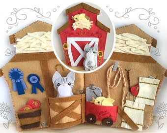 May include: A felt barn play set with a red roof, a brown base, and a gray cat peeking out of a window. The barn has a hayloft, a blue ribbon, a red wagon, and a bucket with apples.