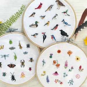 Op de afbeelding: Drie borduurringen met verschillende natuurthema's. De eerste ring heeft een verscheidenheid aan insecten, waaronder vlinders, libellen en kevers. De tweede ring heeft een verscheidenheid aan vogels, waaronder kardinaals, blauwe gaaien en kolibries. De derde ring heeft een verscheidenheid aan bloemen, waaronder klaprozen, madeliefjes en wilde bloemen.