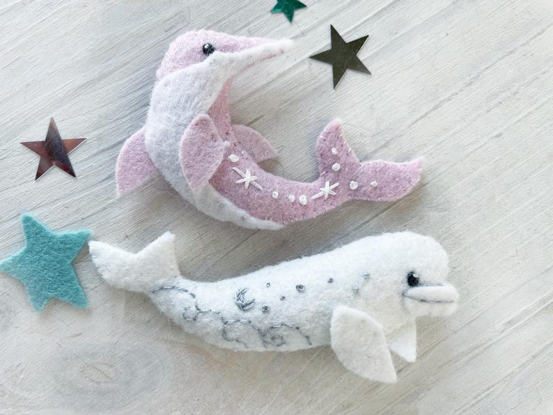 May include: Two felt dolphin ornaments, one pink with white stars and one white with gray stars. The ornaments are on a white wooden background.