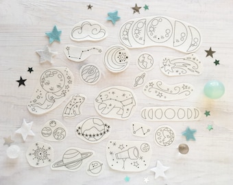 May include: A set of 18 white felt cutouts featuring celestial designs, including moons, stars, planets, and a cloud. The cutouts are arranged on a white wooden surface with blue and silver glitter.
