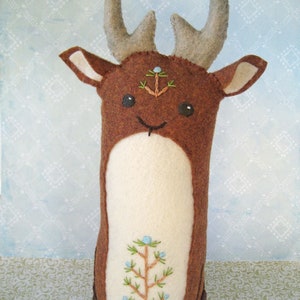 May include: A brown felt deer with a white felt belly and embroidered details. The deer has a small pine tree embroidered on its forehead and a larger pine tree embroidered on its belly. The deer has a stitched smile and large black eyes.