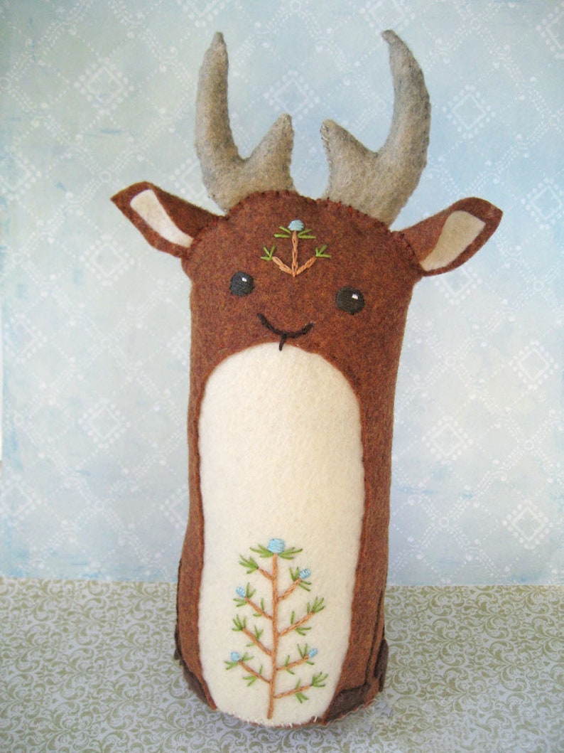 May include: A brown felt deer with a white felt belly and embroidered details. The deer has a small pine tree embroidered on its forehead and a larger pine tree embroidered on its belly. The deer has a stitched smile and large black eyes.