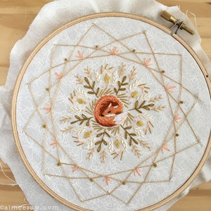 May include: Embroidered hoop art featuring a sleeping fox in shades of orange and white, surrounded by floral and geometric designs. The embroidery is set on a white fabric with a lace pattern, framed by a wooden hoop. The design includes gold and pink accents.