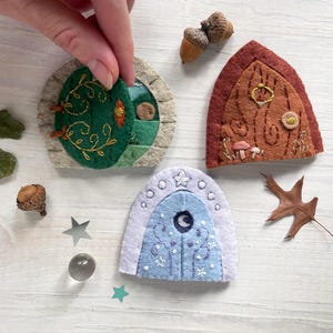 May include: Three felt fairy doors with different designs. The first door is green with a leafy design and a small opening. The second door is brown with a woodgrain design and a gold ring handle. The third door is blue with a moon and stars design.