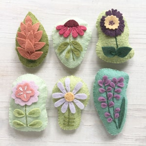 Op de afbeelding: Zes vilten bloem- en bladerenapplicaties in verschillende kleuren en ontwerpen. De applicaties bevinden zich op een witte houten achtergrond.