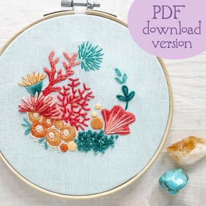 May include: A close-up of a blue embroidery hoop with a colorful floral design. The design features coral, orange, yellow, and teal thread. The hoop is on a white wooden surface with two crystals.