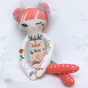 May include: A handmade doll with pink hair and a white dress. The doll has embroidered tattoos on its arms and chest, including a heart, flowers, a skull, and the word "LOVE".