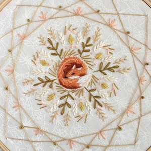 May include: Embroidered hoop art featuring a sleeping fox in shades of orange, brown, and white, surrounded by floral and leaf embroidery in muted tones. The design is set against a white fabric background with a delicate pattern, framed by a wooden hoop.