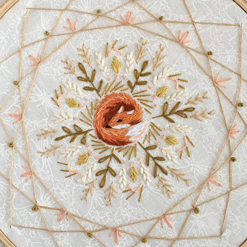 May include: Embroidered hoop art featuring a sleeping fox in shades of orange, brown, and white, surrounded by floral and leaf embroidery in muted tones. The design is set against a white fabric background with a delicate pattern, framed by a wooden hoop.