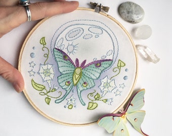 May include: Embroidered hoop art featuring a green and purple luna moth with a white moon and green vines on a white background.