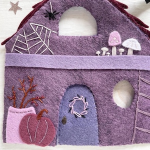 May include: A felt Halloween house in shades of purple, featuring a spiderweb, mushrooms, and a pumpkin. The house has a door with a wreath and a small window. The roof is dark red.