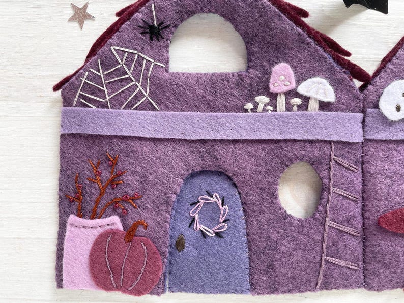 May include: A felt Halloween house in shades of purple, featuring a spiderweb, mushrooms, and a pumpkin. The house has a door with a wreath and a small window. The roof is dark red.