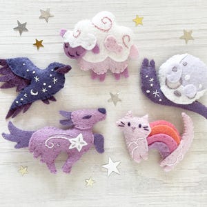 Puede incluir: Una colección de criaturas celestiales de fieltro, que incluye un pájaro morado, una oveja, un caracol con una luna, un unicornio y un gato con una cola de arcoíris. Las figuras son en tonos morado, rosa y blanco, con estrellas.