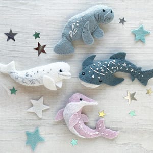 May include: Four felt sea animal toys: a blue manatee, a white beluga whale, a blue whale shark, and a pink dolphin. The toys are arranged on a white wooden surface with silver and blue star-shaped embellishments.