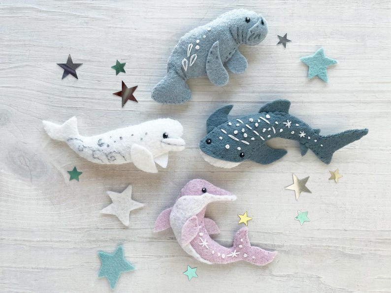 May include: Four felt sea animal toys: a blue manatee, a white beluga whale, a blue whale shark, and a pink dolphin. The toys are arranged on a white wooden surface with silver and blue star-shaped embellishments.
